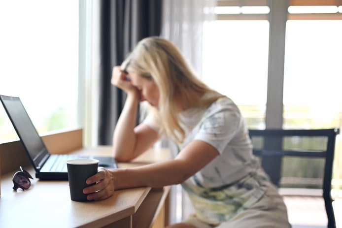 Женщина сидит у окна с чашкой, выглядит уставшей, но спокойной.